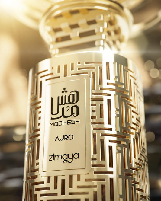 Gold-patterned perfume bottle with 'Modhesh Aura' and 'Zimaya' branding on a blurred background