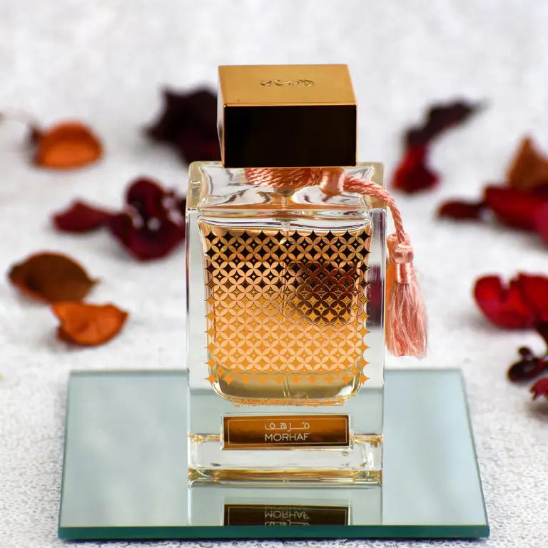 Perfume bottle with gold cap and intricate design on a reflective surface with dried leaves in the background