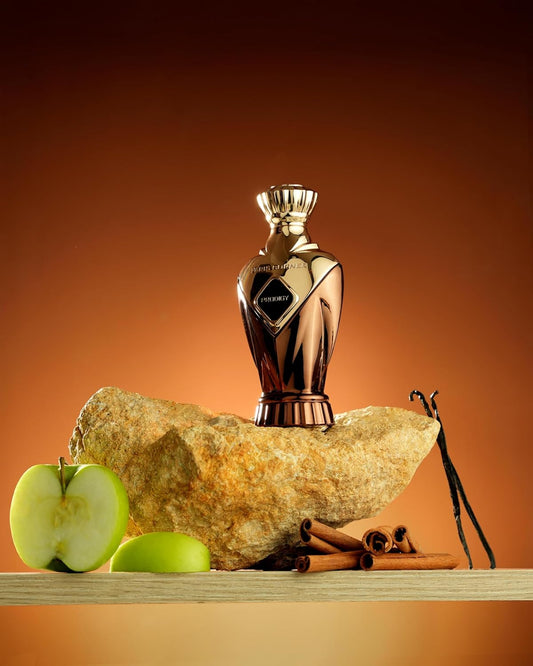 Paris Corner Prodigy perfume bottle on a rock with apples, vanilla beans, and cinnamon sticks on an orange background