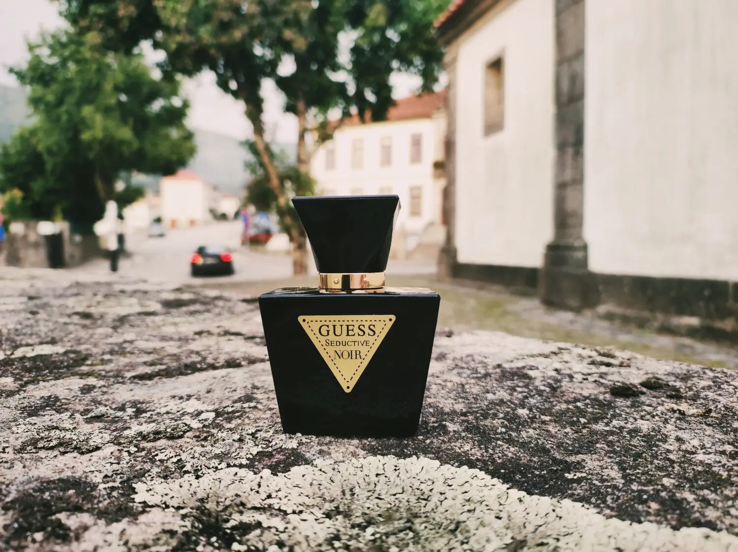 Black Guess perfume bottle on a textured surface with a blurred street scene in the background