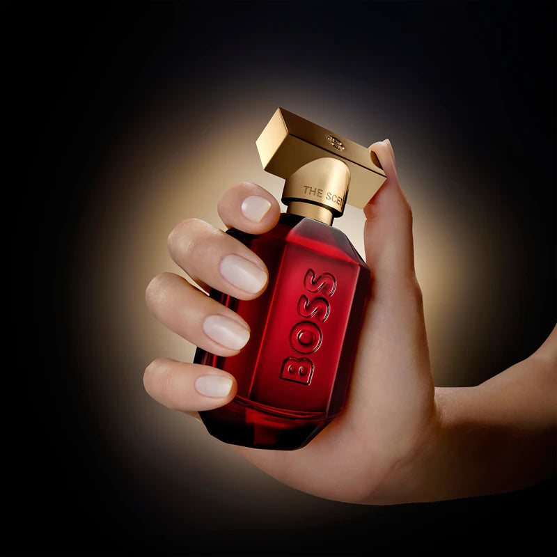 Hand holding a red BOSS perfume bottle with a gold cap against a dark background