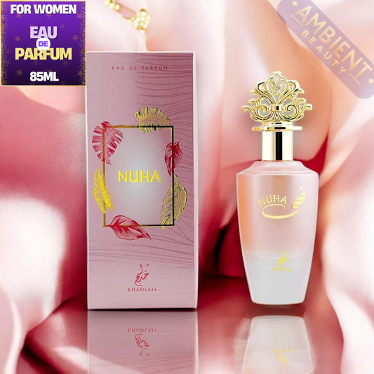 Nuha Eau de Parfum 85ml bottle with its matching pink box, displayed on a reflective surface with draped pink satin fabric. The bottle has a frosted pink body with a shiny gold cap, and the box features pink and gold tropical leaf designs.