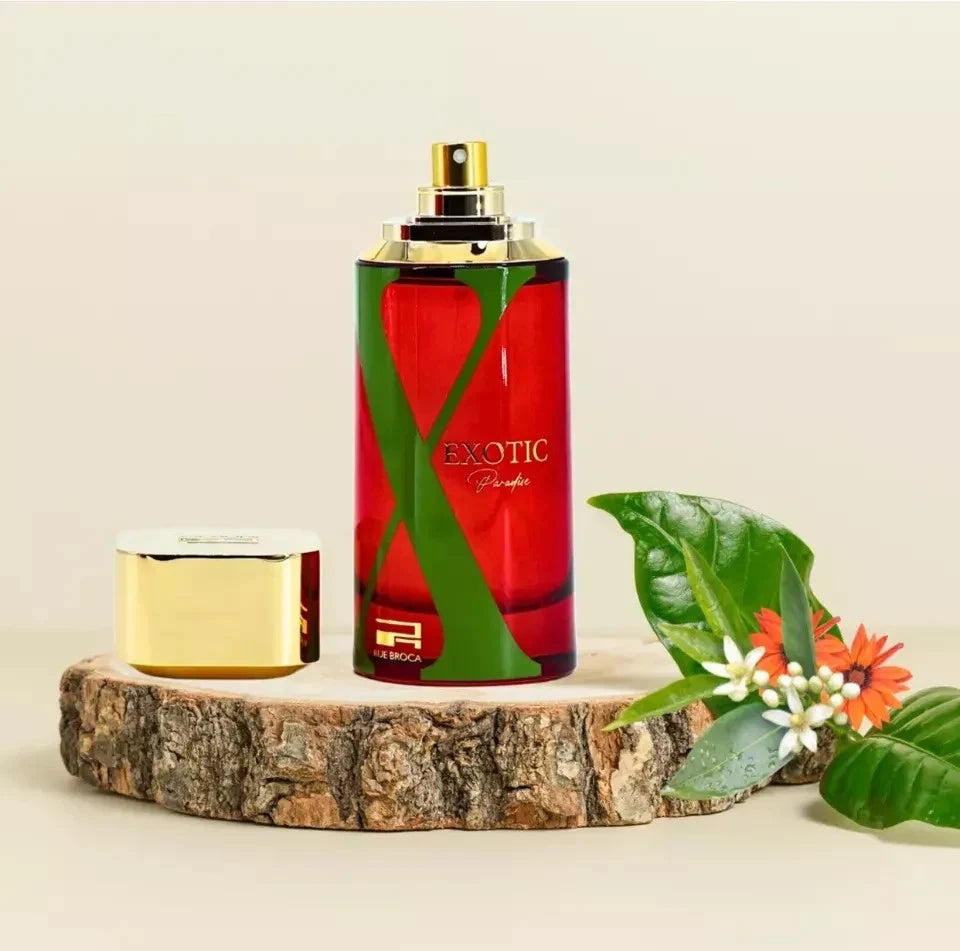 Rue Broca Exotic Paradise Red perfume bottle with gold cap on a wooden platform, surrounded by green leaves and flowers.