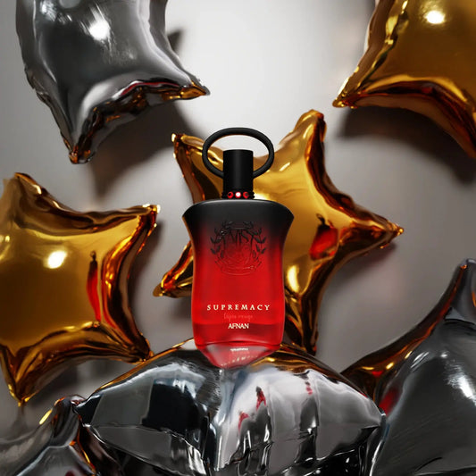 Red perfume bottle labeled 'Supremacy' surrounded by metallic star-shaped objects on a reflective surface.